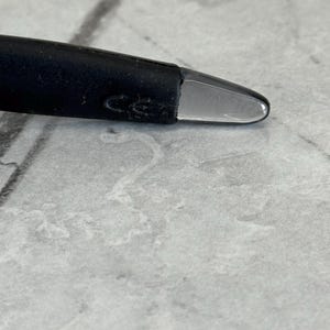 May include: Close-up of a black pen with a clear, rounded tip. The pen's body is dark and textured, with a slight curve. The tip is smooth and transparent, reflecting light. The background is a light gray, textured surface.