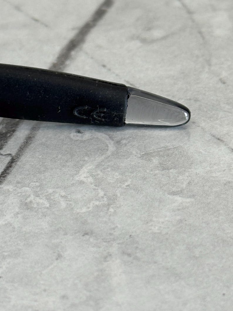 May include: Close-up of a black pen with a clear, rounded tip. The pen's body is dark and textured, with a slight curve. The tip is smooth and transparent, reflecting light. The background is a light gray, textured surface.