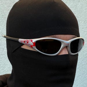 May include: Silver-framed sunglasses with black lenses and red and white accents. The sunglasses have a sporty, streamlined design. The image shows the sunglasses worn with a black balaclava, suggesting a focus on eye protection and style.
