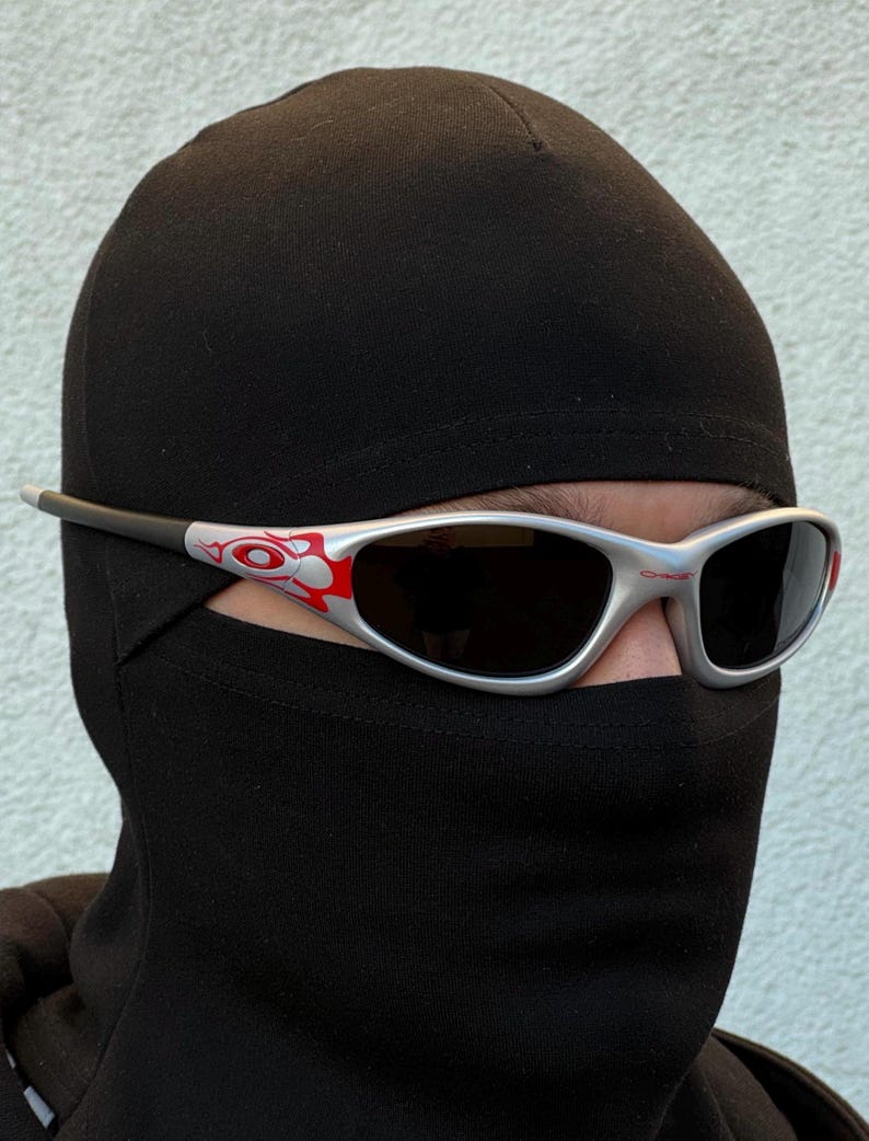 May include: Silver-framed sunglasses with black lenses and red and white accents. The sunglasses have a sporty, streamlined design. The image shows the sunglasses worn with a black balaclava, suggesting a focus on eye protection and style.