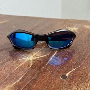 Op de afbeelding: Zwarte zonnebril met blauw gespiegelde glazen. De zonnebril heeft een slank, gebogen montuurontwerp. De glazen reflecteren een heldere blauwe kleur. De zonnebril ligt op een houten oppervlak.