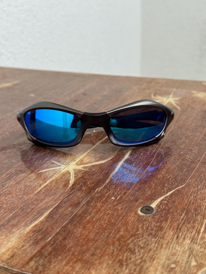 Op de afbeelding: Zwarte zonnebril met blauw gespiegelde glazen. De zonnebril heeft een slank, gebogen montuurontwerp. De glazen reflecteren een heldere blauwe kleur. De zonnebril ligt op een houten oppervlak.