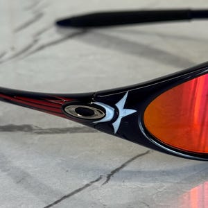 May include: Close-up of black sunglasses with red lenses. The frame has a red and white design on the temples, with a white star logo. The lenses reflect a bright orange color.