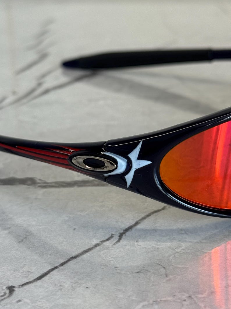 May include: Close-up of black sunglasses with red lenses. The frame has a red and white design on the temples, with a white star logo. The lenses reflect a bright orange color.