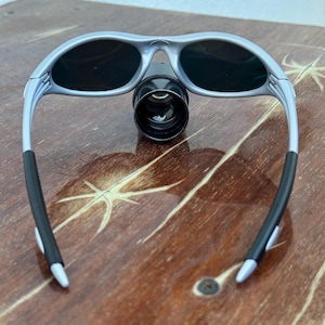 May include: Silver-framed sunglasses with dark lenses and a black cylindrical attachment. The sunglasses have black rubber grips on the temples and are resting on a wooden surface.