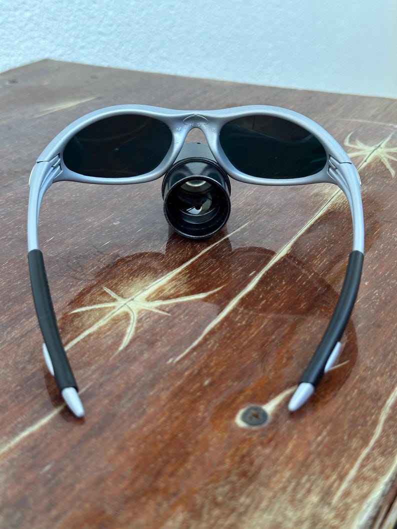 May include: Silver-framed sunglasses with dark lenses and a black cylindrical attachment. The sunglasses have black rubber grips on the temples and are resting on a wooden surface.