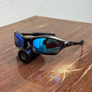 Op de afbeelding: Zwarte zonnebril met blauw gespiegelde glazen. De zonnebril heeft een strak, modern design met een zwart montuur en zwarte pootjes. De glazen reflecteren een helderblauwe kleur. De zonnebril ligt op een zwart cilindrisch object.