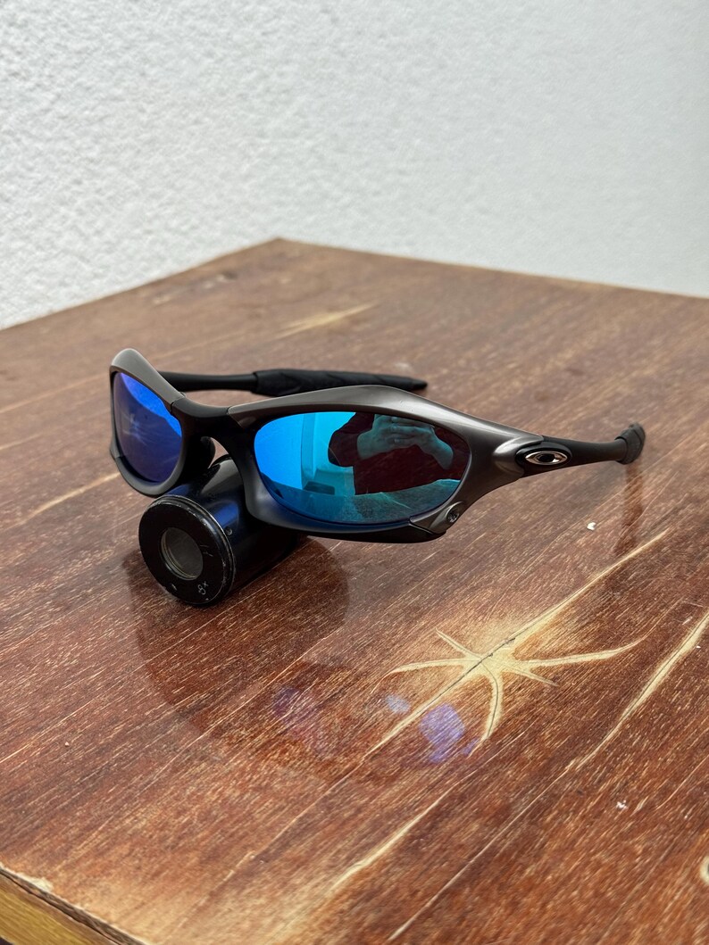 Op de afbeelding: Zwarte zonnebril met blauw gespiegelde glazen. De zonnebril heeft een strak, modern design met een zwart montuur en zwarte pootjes. De glazen reflecteren een helderblauwe kleur. De zonnebril ligt op een zwart cilindrisch object.
