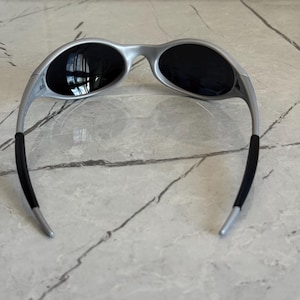 Puede incluir: Gafas de sol plateadas con lentes oscuras. Las gafas de sol tienen un dise&ntilde;o moderno con patillas curvas y un acabado mate. Las lentes son ovaladas y ofrecen cobertura total. Las patillas tienen puntas de goma negra para mayor comodidad.