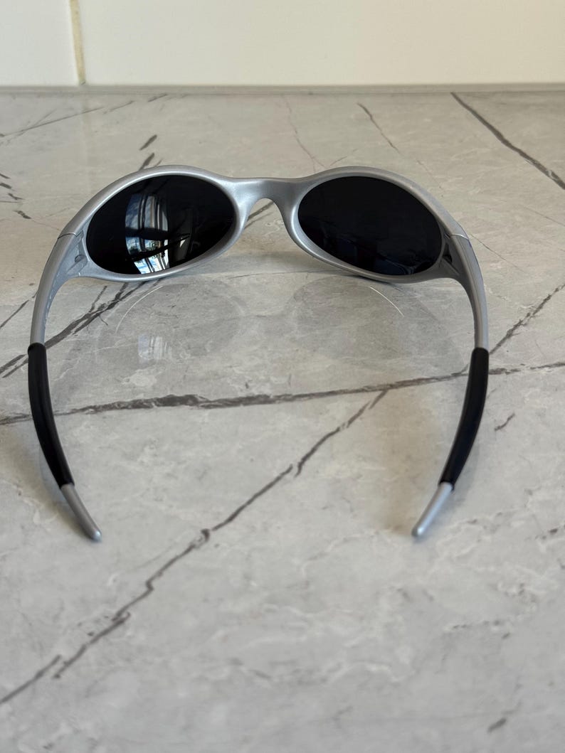 Puede incluir: Gafas de sol plateadas con lentes oscuras. Las gafas de sol tienen un dise&ntilde;o moderno con patillas curvas y un acabado mate. Las lentes son ovaladas y ofrecen cobertura total. Las patillas tienen puntas de goma negra para mayor comodidad.