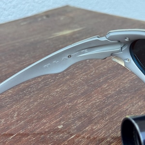 Puede incluir: Primer plano de unas gafas de sol plateadas. Las gafas de sol tienen un dise&ntilde;o elegante y moderno con lentes oscuras. La montura presenta el nombre de la marca. La imagen est&aacute; tomada desde un &aacute;ngulo lateral, mostrando los detalles de la montura y las bisagras.