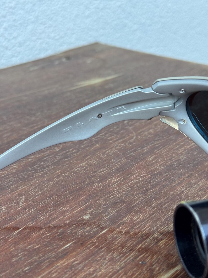 Puede incluir: Primer plano de unas gafas de sol plateadas. Las gafas de sol tienen un dise&ntilde;o elegante y moderno con lentes oscuras. La montura presenta el nombre de la marca. La imagen est&aacute; tomada desde un &aacute;ngulo lateral, mostrando los detalles de la montura y las bisagras.