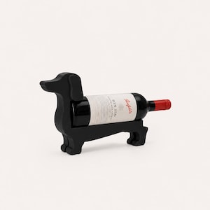 Op de afbeelding: Een matzwarte houten wijnflessenhouder in de vorm van een teckel. De houder heeft een uitsparing in het midden om een wijnfles stevig vast te houden. Een fles Penfolds Bin 150 wijn met een wit etiket en een rode dop is in de houder geplaatst.