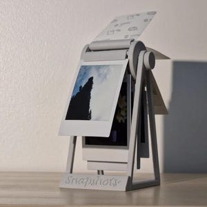 May include: A light gray rotating photo display stand with a triangular frame and the word Snapshots engraved on the base. The stand holds multiple square instant photos, including one showing a dark tree silhouette against a cloudy sky.