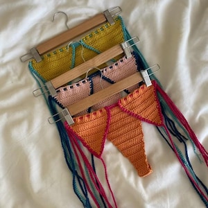Puede incluir: Cuatro tops de bikini de ganchillo hechos a mano en amarillo, rosa y naranja, cada uno en una percha de madera. Los tops tienen adornos coloridos y lazos largos colgantes. El trabajo de ganchillo es visible, mostrando la textura y la artesanía de los trajes de baño.