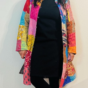 May include: A long, colorful patchwork jacket with a button closure. The jacket is made of different fabrics and patterns, including floral, geometric, and paisley designs. The jacket is worn over a black mock neck shirt and black leggings.