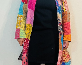 May include: A long, colorful patchwork jacket with a button closure. The jacket is made of different fabrics and patterns, including floral, geometric, and paisley designs. The jacket is worn over a black mock neck shirt and black leggings.
