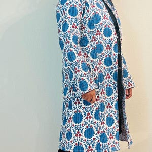 Puede incluir: Una chaqueta larga acolchada blanca con un estampado floral azul y rojo. La chaqueta tiene un cuello mandarín y cierre de botón.
