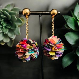 Pode incluir: Um par de brincos de argola com correntes douradas e pingentes coloridos em forma de pompom feitos de lantejoulas.