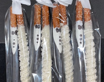 Puede incluir: Palitos de pretzel cubiertos de chocolate blanco y negro, decorados para parecer esmóquines y vestidos de novia. Cada pretzel está envuelto individualmente en celofán y atado con una cinta blanca. Un dulce para bodas.