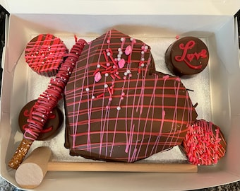 May include: A heart-shaped chocolate cake decorated with pink and red sprinkles and chocolate drizzle. The cake is surrounded by smaller chocolate treats, including a pretzel rod dipped in chocolate and sprinkles, and a chocolate disc with the word "Love" written on it.
