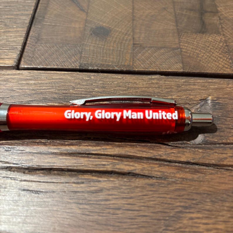 May include: A red pen with a silver clip and tip. The pen has the text "Glory, Glory Man United" printed in white. The pen is lying on a wooden surface.