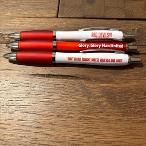 May include: Three ballpoint pens with red and white barrels. The pens have silver accents and text that reads "RED DEVILS!!!", "Glory, Glory Man United", and "DON'T GO OUT TONIGHT, UNLESS YOUR RED AND WHITE". The pens are on a wooden surface.