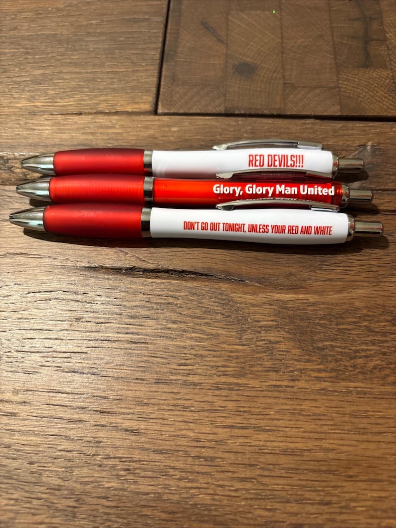 May include: Three ballpoint pens with red and white barrels. The pens have silver accents and text that reads "RED DEVILS!!!", "Glory, Glory Man United", and "DON'T GO OUT TONIGHT, UNLESS YOUR RED AND WHITE". The pens are on a wooden surface.