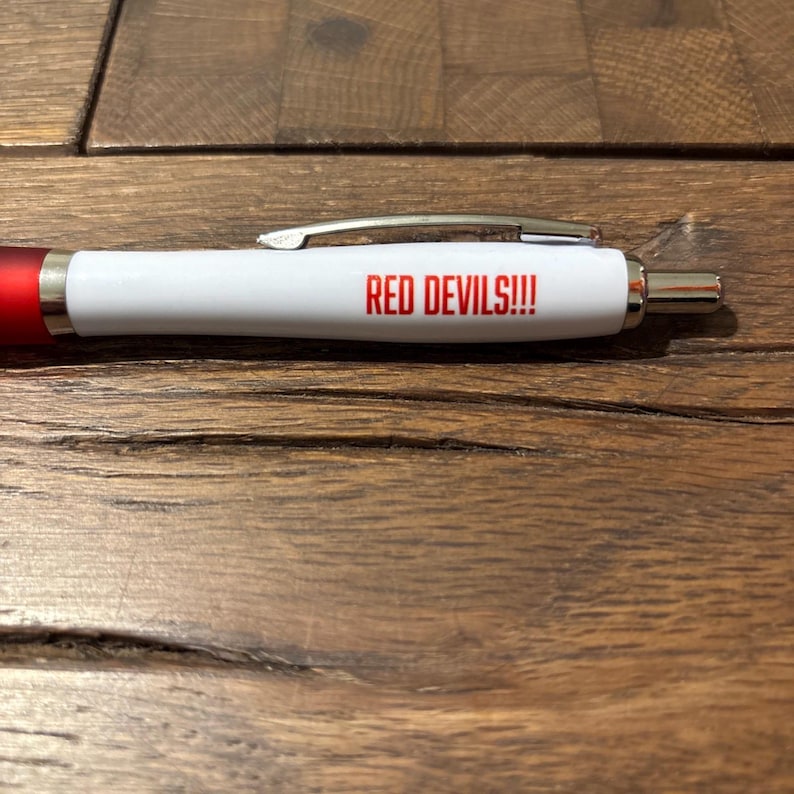 May include: A white pen with a red tip and the text "RED DEVILS!!!" printed in red. The pen has a silver clip and a silver tip. The pen is on a wooden surface.