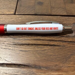 May include: A white pen with red accents and silver clip. The pen has the text "DON'T GO OUT TONIGHT, UNLESS YOUR RED AND WHITE" printed in red. The pen is lying on a wooden surface.