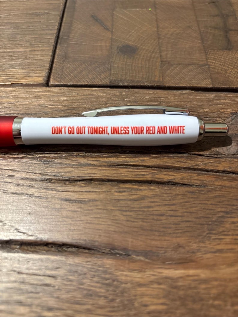 May include: A white pen with red accents and silver clip. The pen has the text "DON'T GO OUT TONIGHT, UNLESS YOUR RED AND WHITE" printed in red. The pen is lying on a wooden surface.
