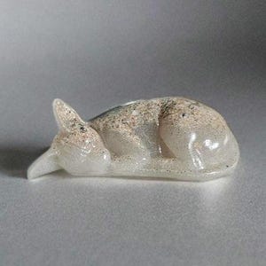 Könnte beinhalten: Eine kleine, schlafende Katzenfigur aus Resin in Weiß mit einer gesprenkelten, hellbraunen Oberfläche.