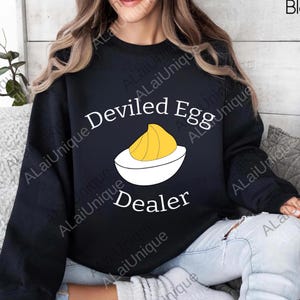 Può includere: Felpa nera con la scritta "Deviled Egg Dealer" e una grafica di un uovo farcito. La grafica presenta una metà di uovo bianco con ripieno giallo. La felpa è uno stile casual e confortevole.