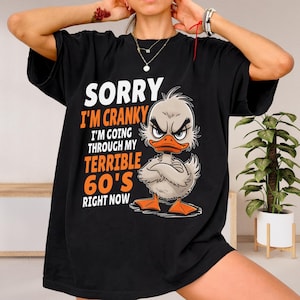 以下が含まれることがあります： 「SORRY I'M CRANKY I'M GOING THROUGH MY TERRIBLE 60'S RIGHT NOW」の文字が入った黒いTシャツ。怒った表情のカモの漫画が描かれています。カモは灰色の羽、オレンジ色のくちばしと足を持っています。
