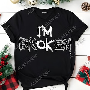 Może przedstawiać: Czarny t-shirt z białym napisem "I'M BROKEN" w stylu distressed. Koszulka ma dekolt w serek i krótkie rękawy. Projekt jest wyśrodkowany z przodu.