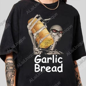 以下が含まれることがあります： サングラスをかけた骸骨がガーリックブレッドを持っている黒いTシャツ。白で「Garlic Bread」の文字がプリントされています。面白くてユニークなデザインです。