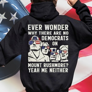 Puede incluir: Sudadera con capucha negra con un gráfico de las figuras del Monte Rushmore con gafas de sol con la bandera estadounidense. El texto dice: "¿Alguna vez te has preguntado por qué no hay demócratas en el Monte Rushmore? Yo tampoco."