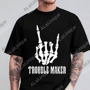 Könnte beinhalten: Schwarzes T-Shirt mit weißem Skelett-Hand-Motiv und dem Text "TROUBLE MAKER" in gotischer Schrift. Das Design ist mittig auf der Vorderseite platziert. Das T-Shirt hat einen Rundhalsausschnitt.
