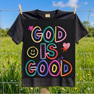 Puede incluir: Camiseta negra con la frase "GOD IS GOOD" en letras coloridas de estilo manuscrito. El diseño incluye una carita sonriente, un corazón y estrellas. La camiseta está colgada en un tendedero frente a un campo verde.