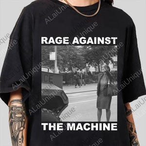 May include: Black t-shirt with the text "RAGE AGAINST THE MACHINE" in white block letters. The shirt features a black and white image of a woman gesturing in front of a police vehicle.