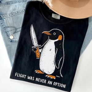Könnte beinhalten: Schwarzes T-Shirt mit einer Pinguin-Illustration, die ein Messer hält. Der Pinguin ist schwarz und weiß mit orangefarbenen Akzenten. Der Text "FLIGHT WAS NEVER AN OPTION" ist unten auf dem Shirt aufgedruckt.