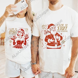 Op de afbeelding: Twee witte T-shirts met kerstthema-afbeeldingen. Eén shirt heeft een retro-illustratie van een vrouw met bellen en de tekst "I love to jingle his bells". Het andere shirt toont de kerstman en de tekst "I love to stuff her stocking."