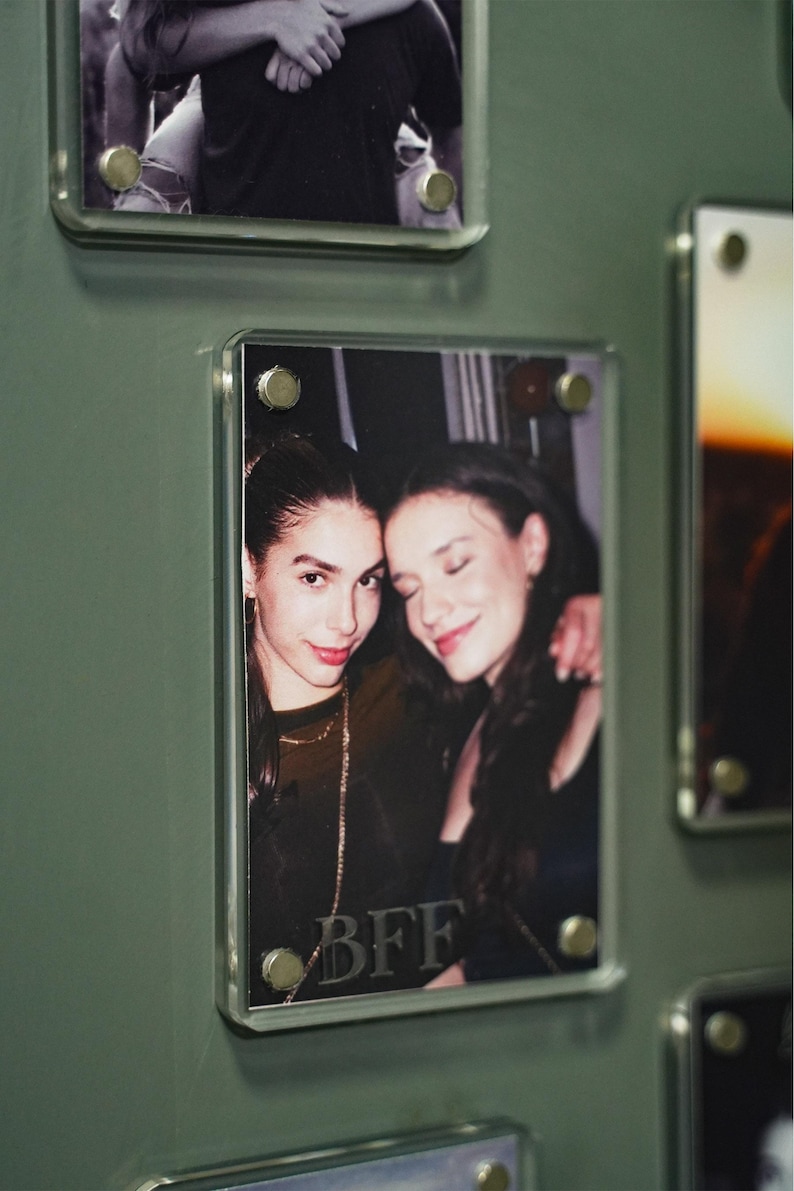 Op de afbeelding: Een verzameling ingelijste foto's aan een groene muur. E&eacute;n frame toont twee lachende vrouwen, met de letters "BFF" zichtbaar. De rechthoekige frames zijn bevestigd met metalen bevestigingsmiddelen.