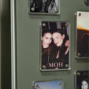 Op de afbeelding: Een verzameling heldere acryl fotomagneten, elk met een andere foto. De foto's tonen twee vrouwen in verschillende poses, waaronder een zwart-wit foto en een zonsondergang. Het woord "MOH" is zichtbaar op een van de foto's.