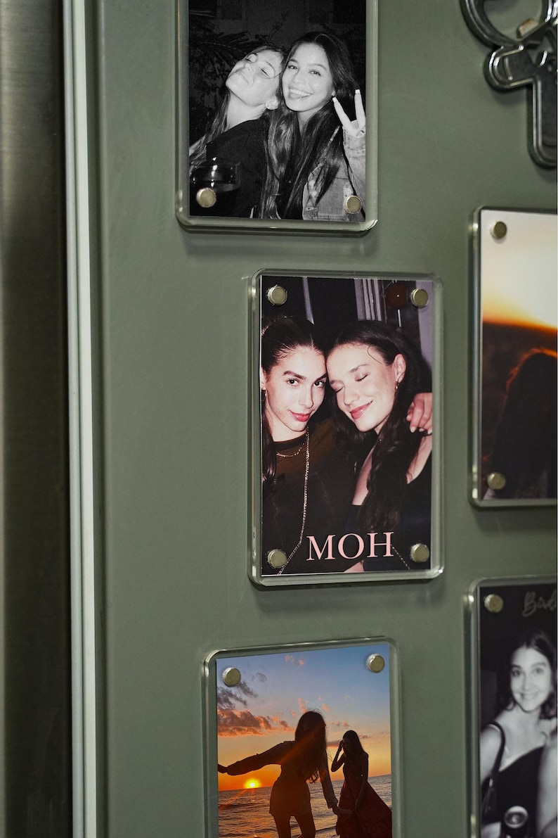 Op de afbeelding: Een verzameling heldere acryl fotomagneten, elk met een andere foto. De foto's tonen twee vrouwen in verschillende poses, waaronder een zwart-wit foto en een zonsondergang. Het woord "MOH" is zichtbaar op een van de foto's.