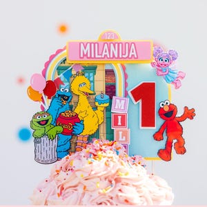May include: A colorful cake topper featuring Sesame Street characters, including Elmo, Big Bird, and Cookie Monster. The topper has the text "CAKE TOPPER" and a name, "MILANIJA", with the number "1". The cake is topped with pink frosting and sprinkles.