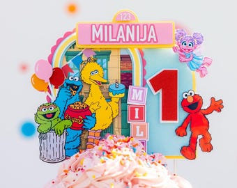 May include: A multi-layered Sesame Street themed cake topper featuring a pink sign with the name MILANIJA, a large red number 1, and illustrations of Elmo, Big Bird, Cookie Monster, Oscar the Grouch, and Abby Cadabby, set against a light blue background.