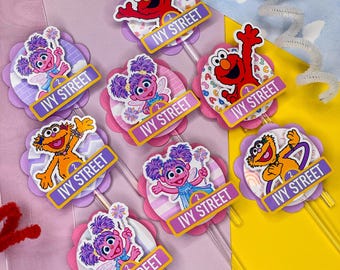May include: A collection of seven decorative cupcake toppers featuring Sesame Street characters Elmo, Abby Cadabby, and Zoe. Each topper includes a purple banner with the text IVY STREET and a small circle with the number 2, mounted on clear sticks.
