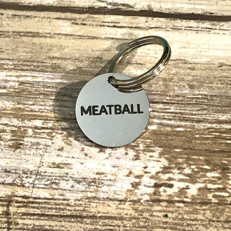 以下が含まれることがあります： 黒い大文字で「MEATBALL」と刻印された、円形の銀色金属製ペット用ネームタグ。タグの上部には、首輪やキーリングに取り付けるための銀色の金属製二重リングが付いています。