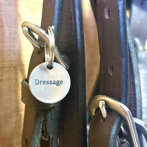 以下が含まれることがあります： Dressageという文字が刻印された円形の銀色の金属タグ。銀色の金属製バックルが付いたダークブラウンのレザーストラップに、二重リングで取り付けられています。タグは滑らかで平らで、中央に鮮明な黒いフォントで文字が記されています。